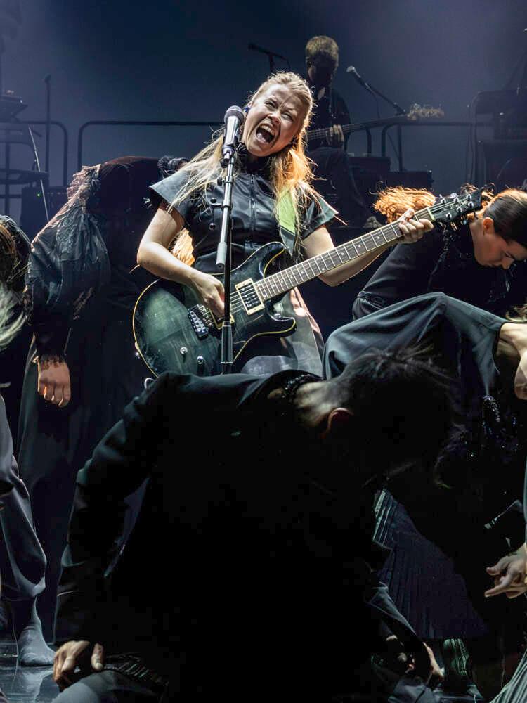 En sångare under scenljus med en elgitarr vrålar i en mikrofon. Runt henne står flera svartklädda dansare med böjda huvuden.