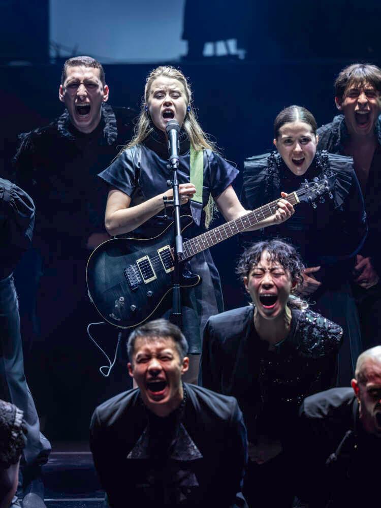 En sångare med elgitarr sjunger i en mikrofon. Hon är omgiven av 10 dansare som också är vända framåt och sjunger högt.