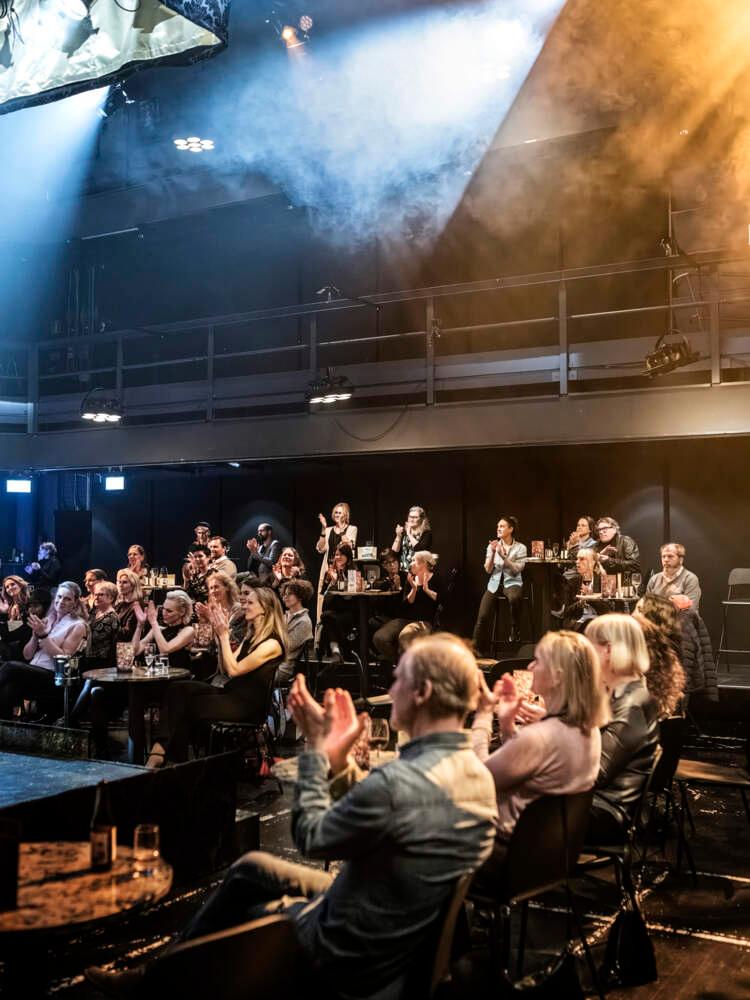 En publik sitter och applåderar i en liten teater, där scenrök syns i strålkastarna som lyser mot scenen.