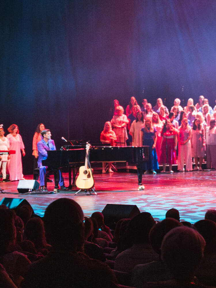 En kör på cirka 100 personer sjunger på en väl upplyst scen i en annars mörk salong. Musiker ackompanjerar dem.