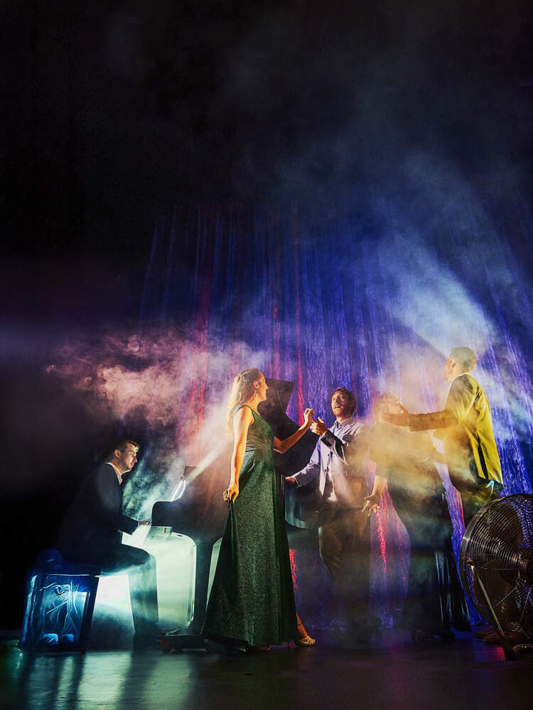 Tre sångare står mittemot varandra  i scenrök och färgade ljus och sjunger framför en ridå. De ackompanjeras av en pianist.