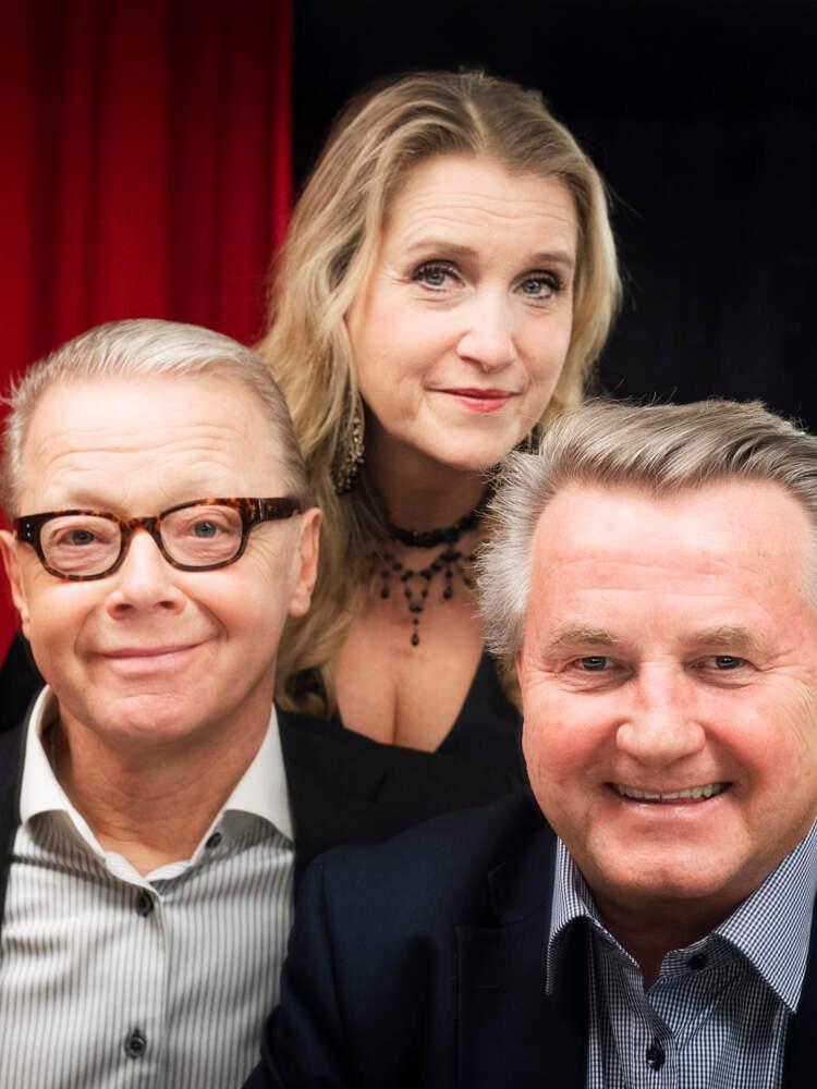 En operasångerska står bakom en annan sångerska och en pianist, med händerna på deras axlar. Alla tittar in i kameran.