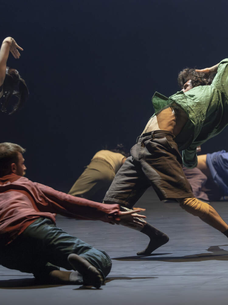 Två dansare står, lutade tillbaka. På golvet runt dem är tre andra dansare på knä. Alla är upplysta ovanifrån.