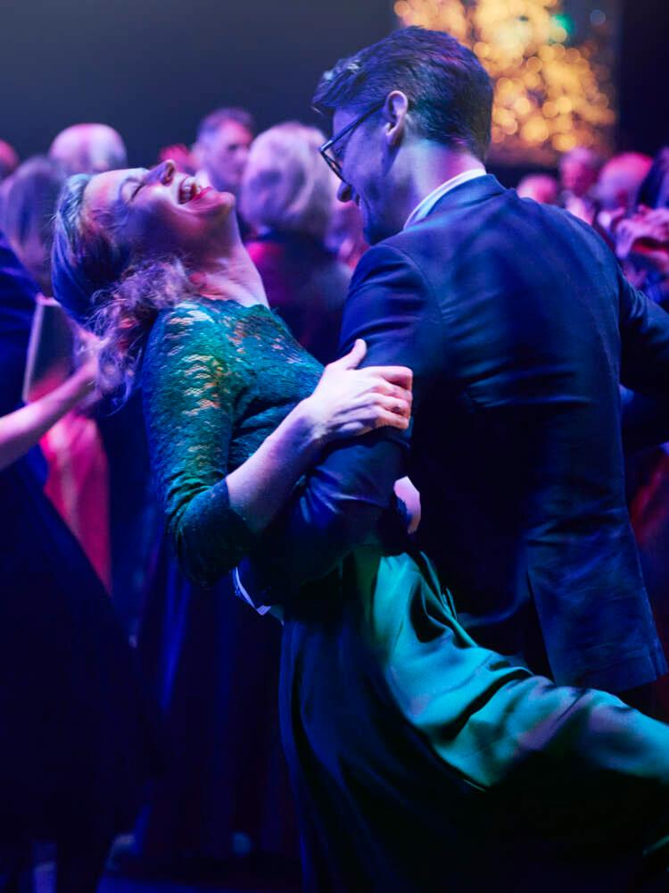 Många par dansar i en balsal under lila ljus. En av paret i förgrunden lutar sig bakåt medan hennes partner håller i henne.