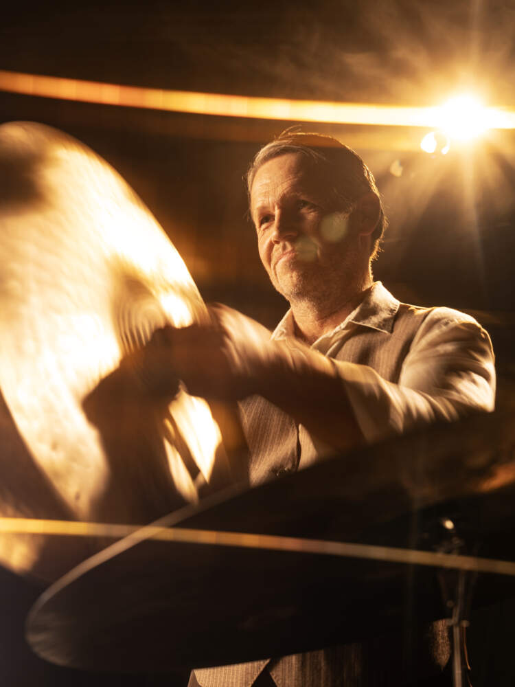 En slagverkare står i gyllene ljus och håller två cymbaler framför sig. Han ler lätt.