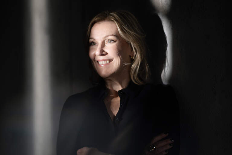 En leende kvinna lutar sig mot en betongvägg med armar i kors. Solljus skiner på hennes ansikte men rummet är annars mörkt.