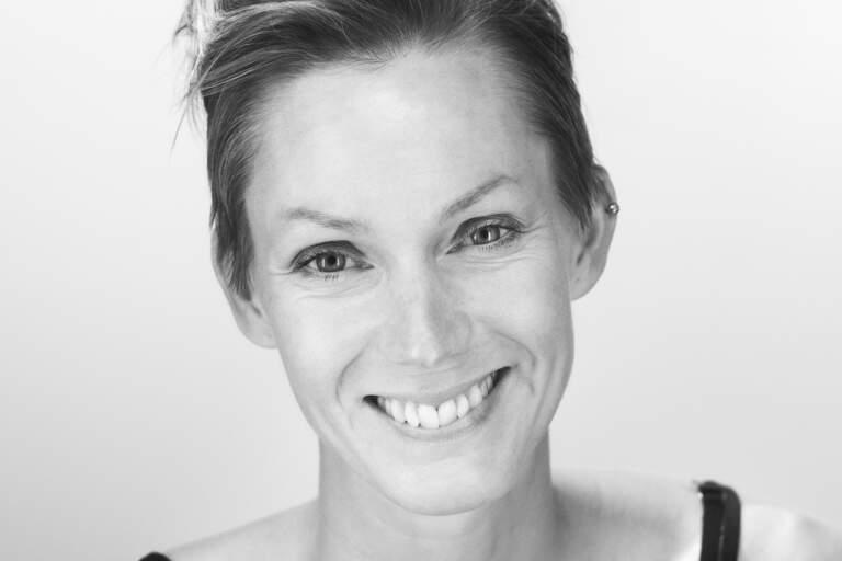En svartvit porträttfoto av en kvinna som sitter framför en ljus bakgrundsfond. Hon tittar in i objektivet.