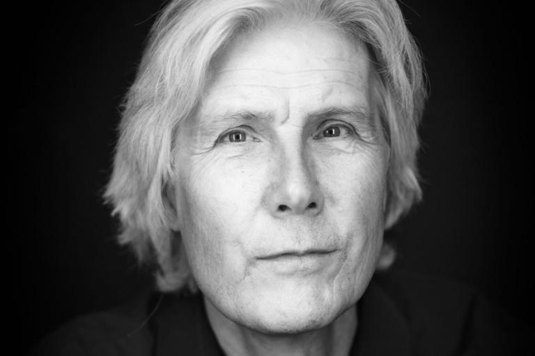 En svartvit bystporträttfoto av en man som sitter framför en svart fond. Han tittar in i objektivet mot betraktaren.