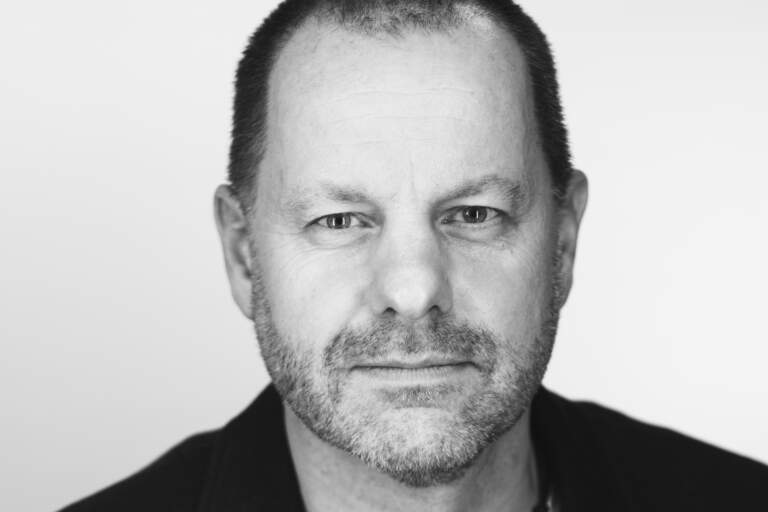 En svartvit bystporträttfoto av en man som sitter framför en ljus fond. Han tittar in i objektivet mot betraktaren. En svartvit bystporträttfoto av en man som sitter framför en ljus bakgrundsfond. Han tittar in i objektivet mot betraktaren.