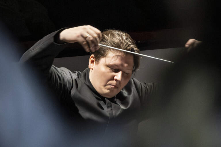 En dirigent, upplyst ovanifrån, står med slutna ögon och armarna upplyfta. Han håller en dirigentpinne i sin högra hand.