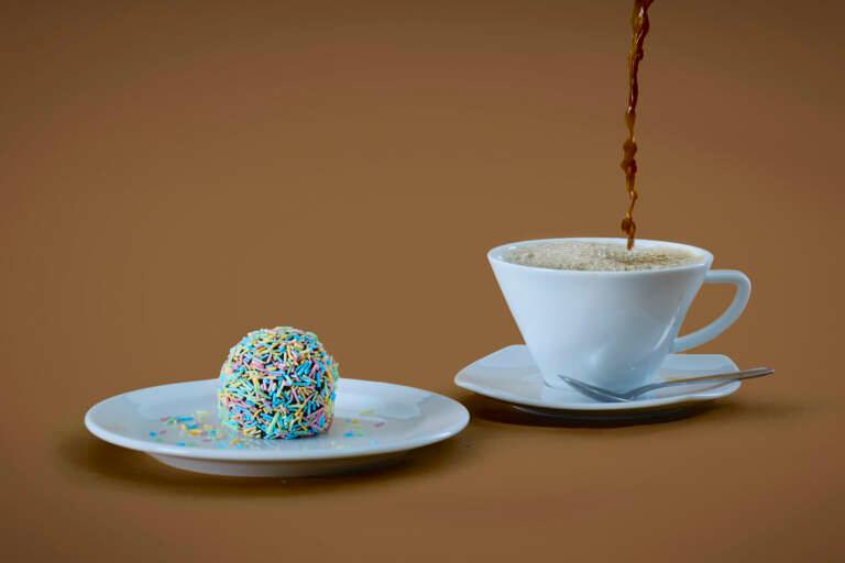 En chokladboll täckt med färgglatt strössel sitter på en assiett bredvid en kaffekopp där kaffet hälls i.