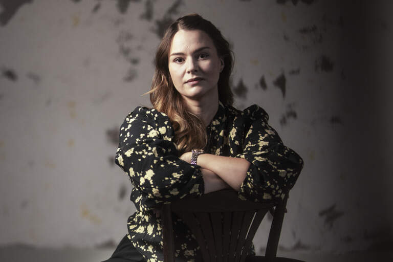 En operasolist sitter på en trästol med underarmarna mot ryggstödet och högerfoten uppdragen på stolen. Hon är barfota.