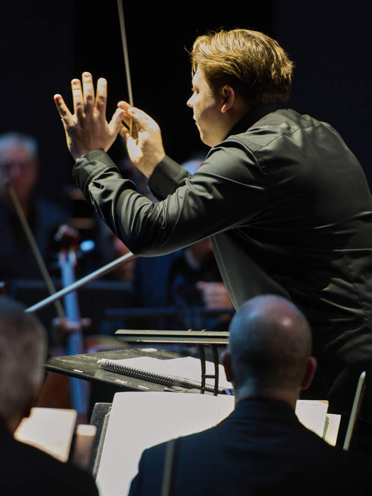 En dirigent står i rampljus och håller upp en öppen handflata. Andra handen håller dirigentpinnen. Han är omgiven av musiker.