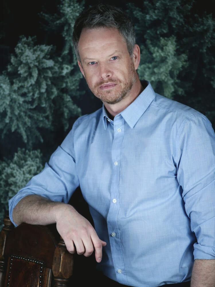 En man står med armen vilande på ryggstödet på en stol av trä och läder. Han bär en ljusblå skjorta med krage.