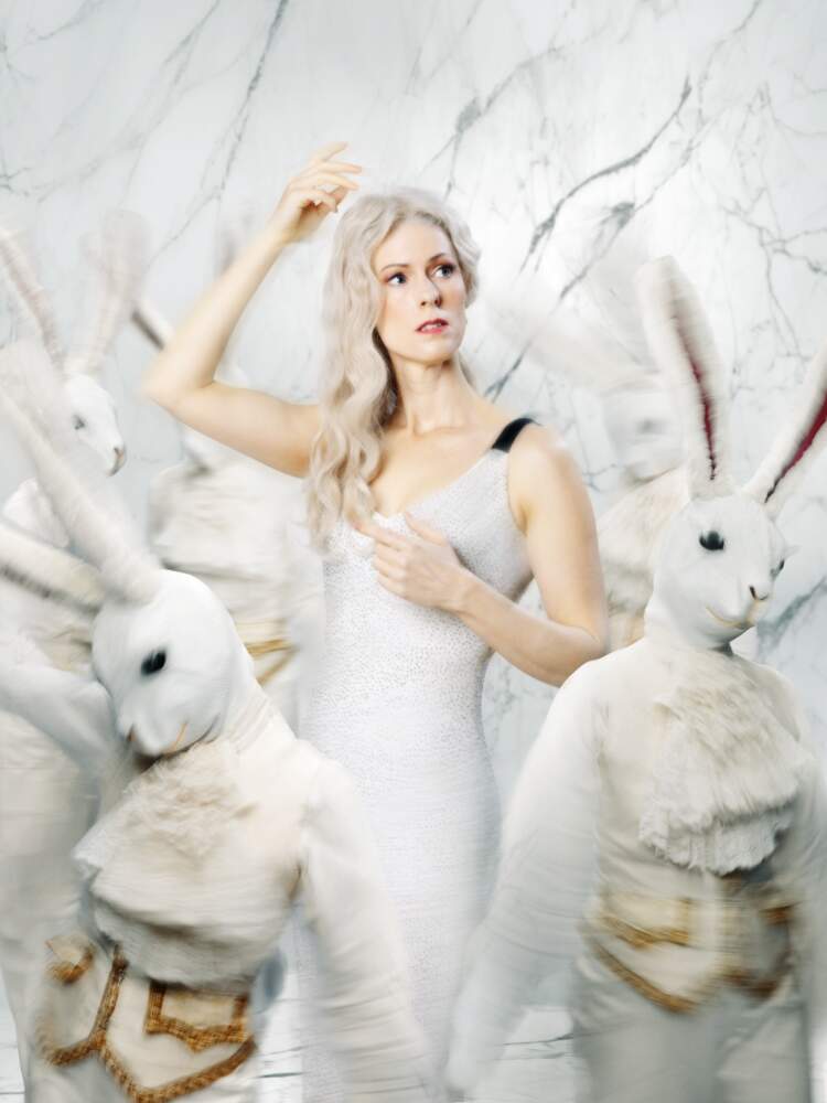 Violetta står i ett vitt marmorrum iklädd en vit klänning. Vita kaniner i guldkantade vita västar och kragar dansar runt.