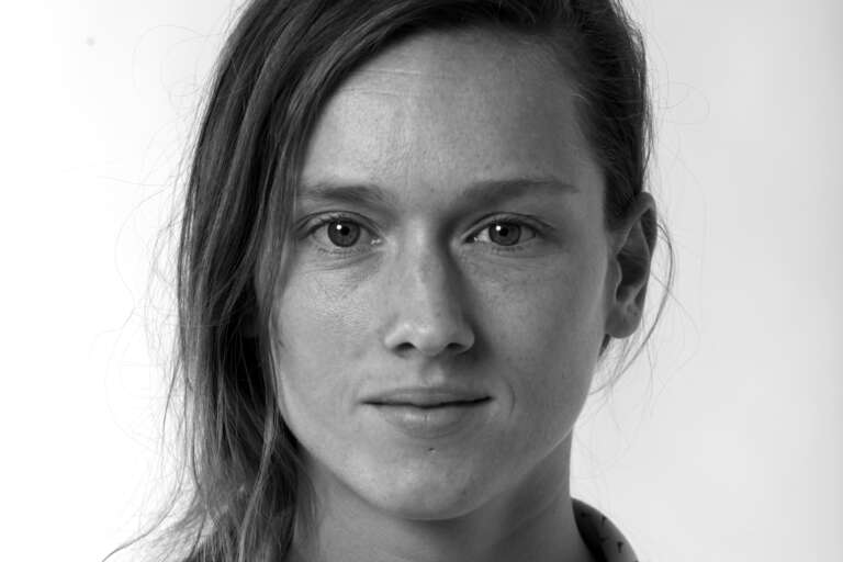 En svartvit porträttfoto av en kvinna som sitter framför en ljus bakgrundsfond. Hon tittar in i objektivet.