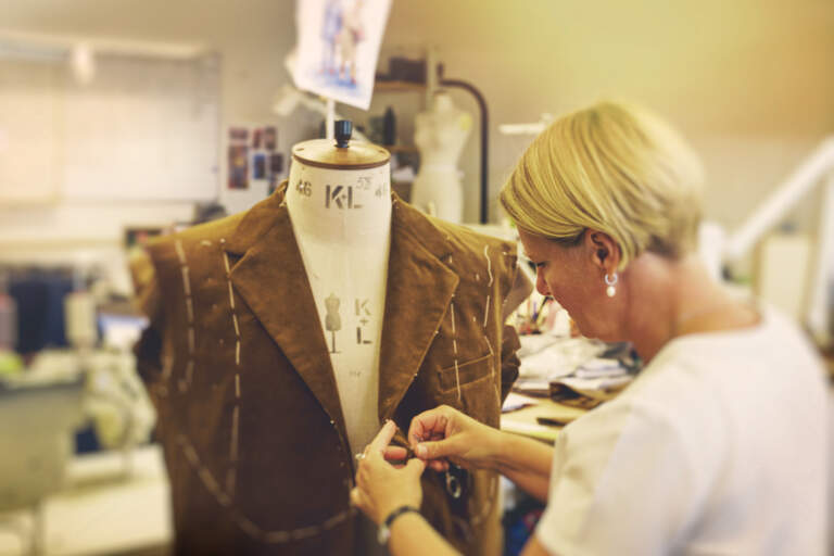 En skräddare står i en ateljé framför en kostymdocka. Hon sätter in en nål nära slaget på en brun kostymjacka.