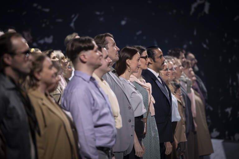 En rad körsångare står leende axel mot axel under publikens applåder i slutet av en föreställning.