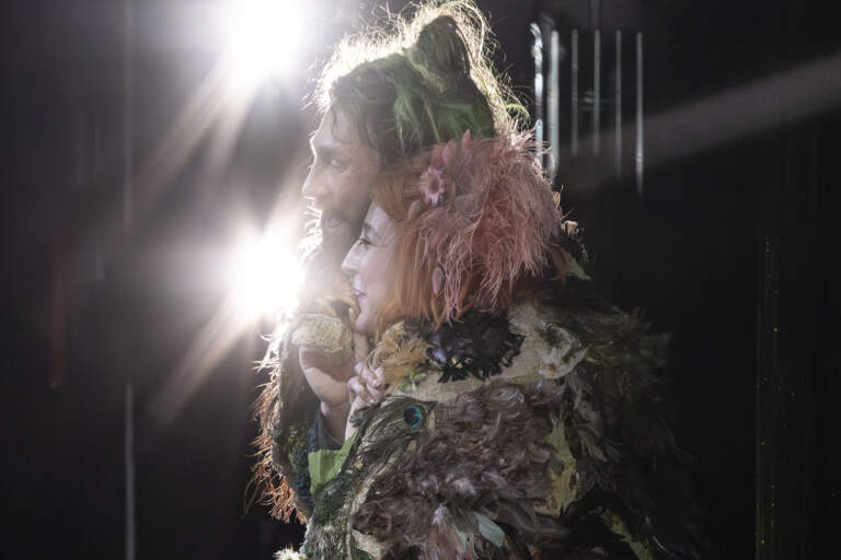 En grönhårig Papageno i fjäderkappa omfamnar en rödhårig Papagena i mossfärgad tunika. De ler.