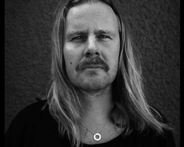 En svartvit bystporträttfoto av en man som sitter framför en svart fond. Han tittar in i objektivet mot betraktaren.