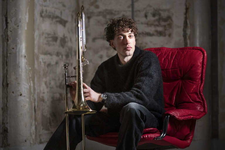 En trombonist sitter i en röd läderfåtölj och håller sitt instrument. Han är barfota och är upplyst från vänster sida.