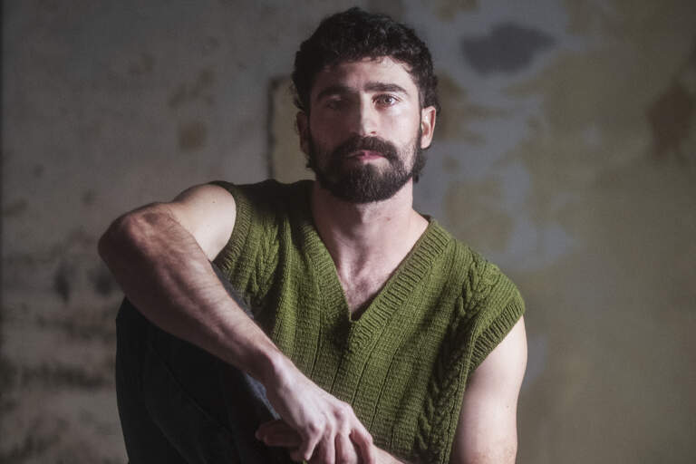 En man sitter barfota på en barstol i ett mörkt rum. Högerknät är uppdraget mot bröstet. Han är upplyst från vänster.