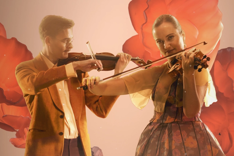 Två violinister står och spelar sina instrument. Stora kronblad liknande former svävar runt dem i persikofärgat ljus.