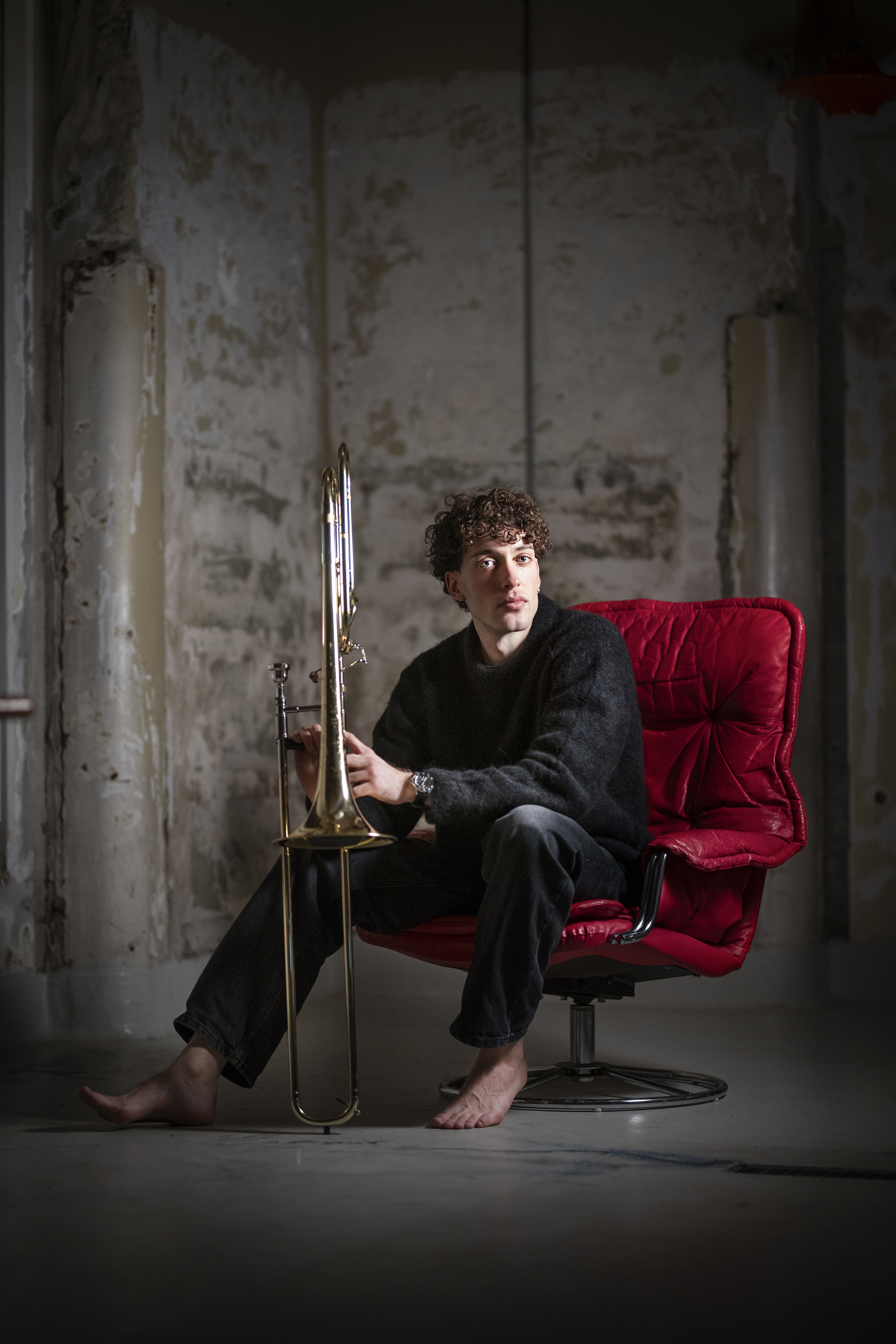 En trombonist sitter i en röd läderfåtölj och håller sitt instrument. Han är barfota och är upplyst från vänster sida.