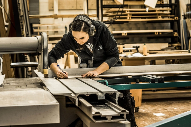En snickare med hörselskydd markerar en bräda med en penna. Brädan vilar på en industriell kapsåg.