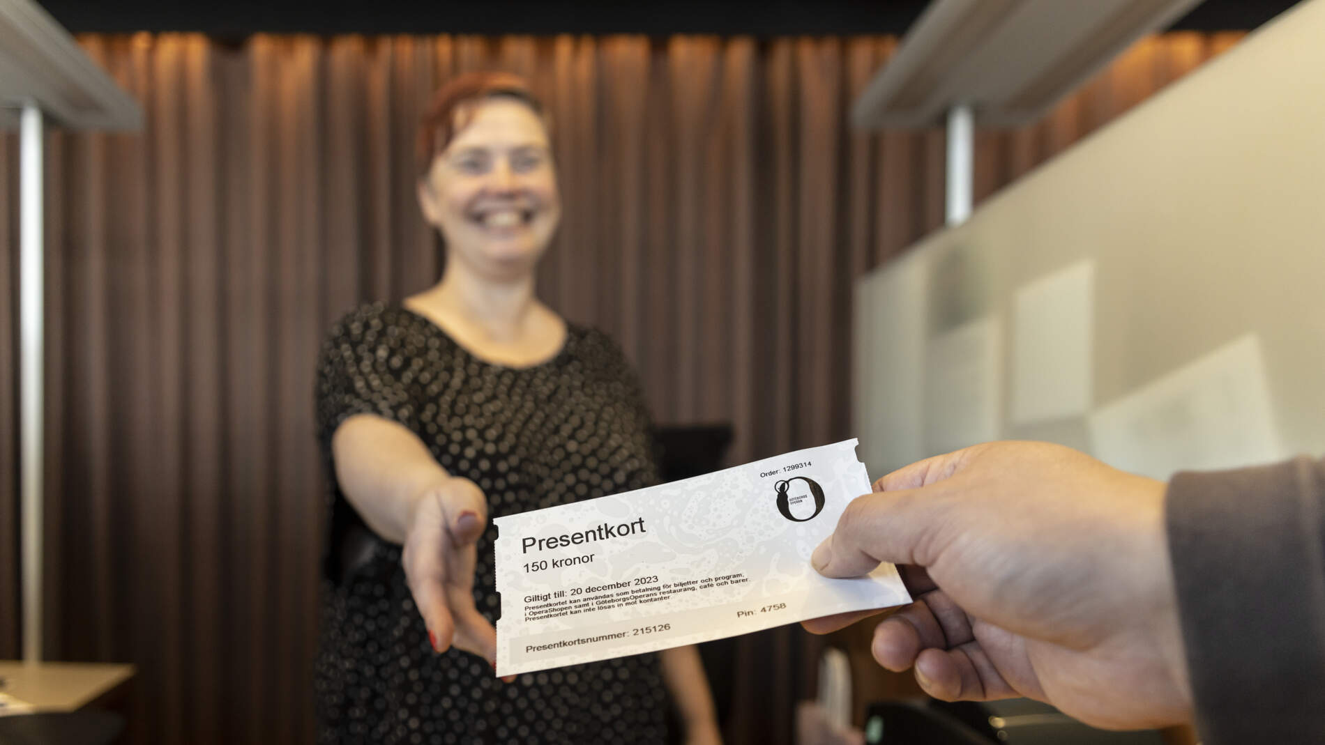 Närbild på en hand som räcker fram ett presentkort till ett leende person bakom disken i en biljettkassa.