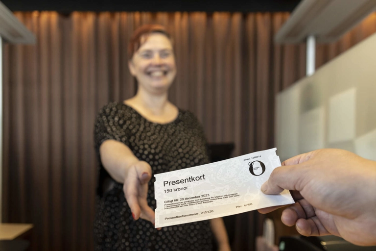 Närbild på en hand som räcker fram ett presentkort till ett leende person bakom disken i en biljettkassa.
