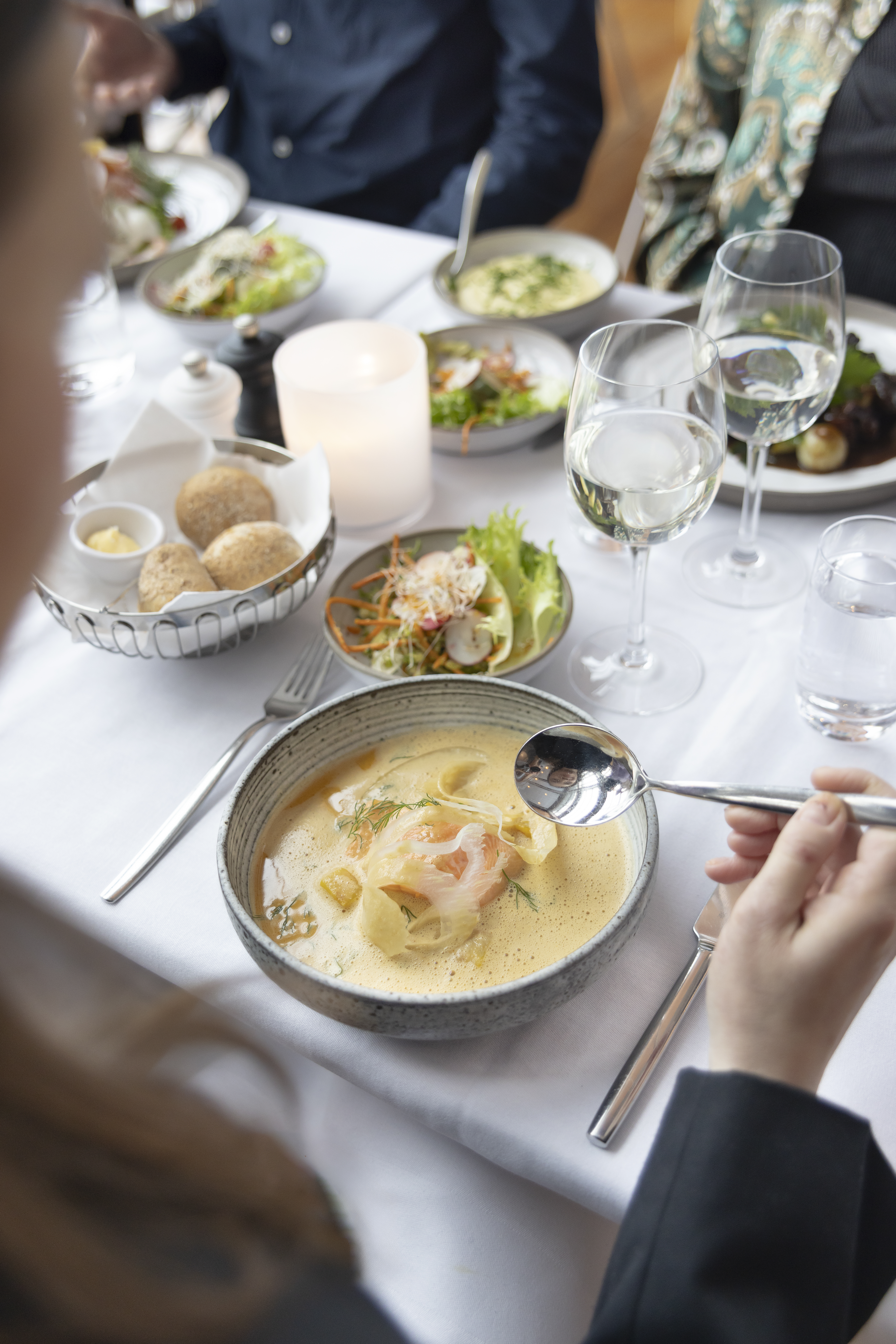 En skål skaldjurssoppa på ett bord, med en sked redo över soppskålen. På bordet står vinglas, sallad och en brödkorg.