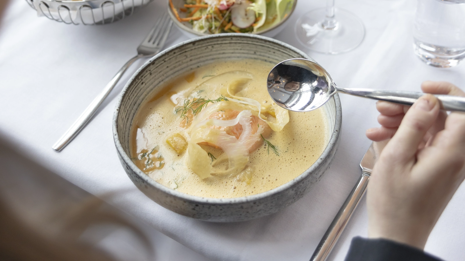 En skål skaldjurssoppa på ett bord, med en sked redo över soppskålen. På bordet står vinglas, sallad och en brödkorg.