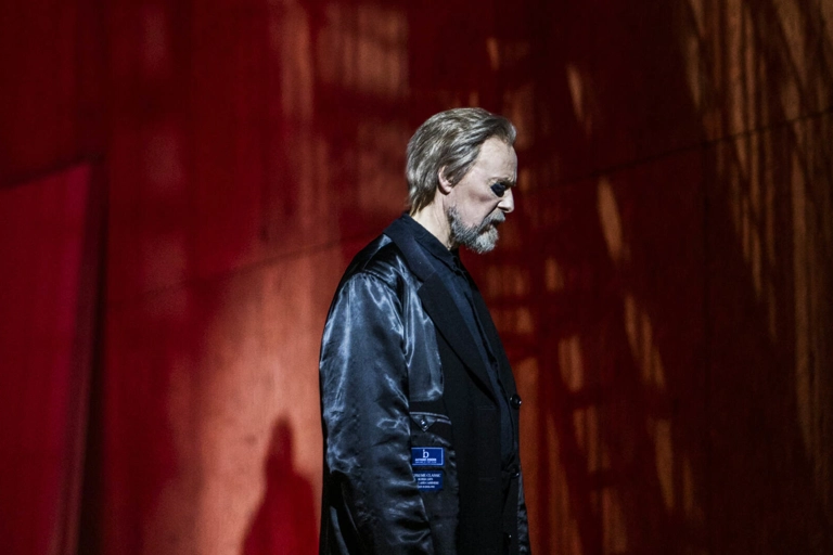 Wotan, med ögonlapp, går med huvudet lätt böjt framför en rödbelyst bakgrund.