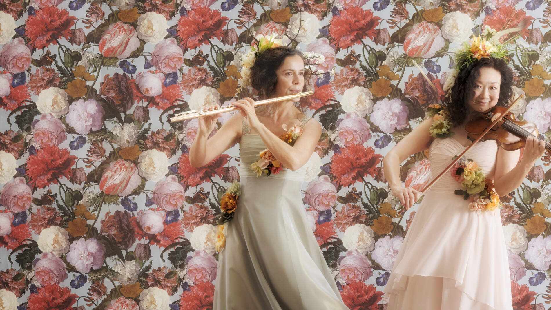 En flöjtist och violinist i långa sidenklänningar och blomsterkransar står framför en blomsterfond och spelar sina instrument.