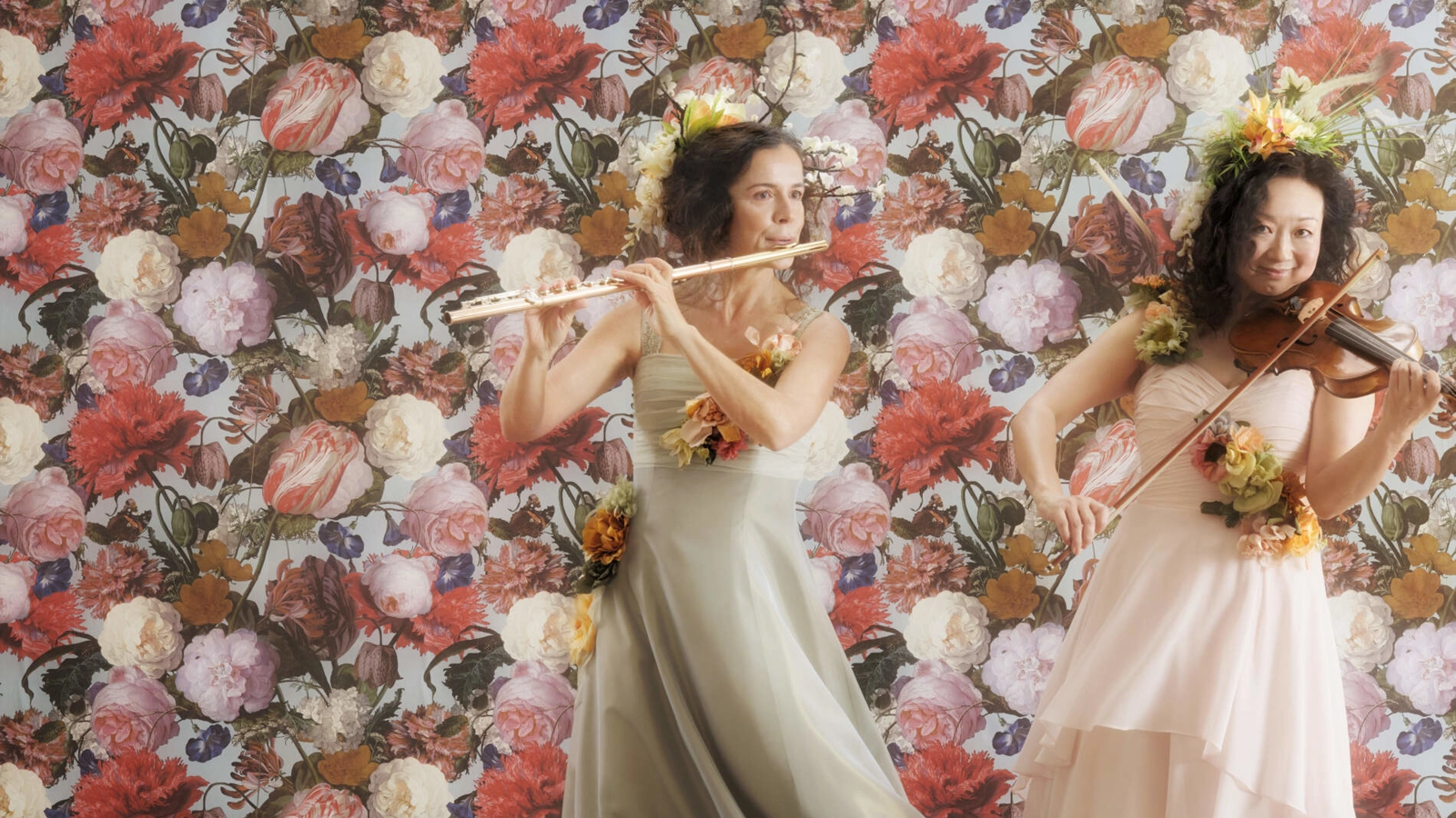 En flöjtist och violinist i långa sidenklänningar och blomsterkransar står framför en blomsterfond och spelar sina instrument.
