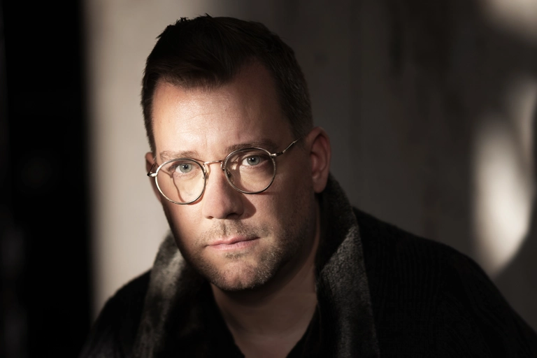 En man sitter framåtlutad. Solljus skiner på ansiktet och delar av betongväggen bakom honom, men rummet är annars i skugga.