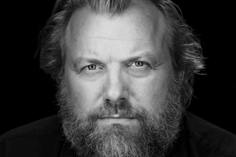 En svartvit bystporträttfoto av en man som sitter framför en svart fond. Han tittar in i objektivet mot betraktaren.