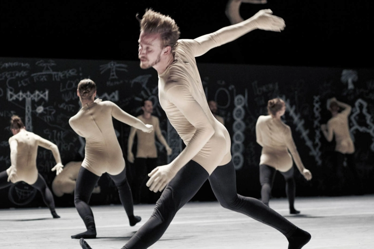 En dansare i beige trikå med svarta strumpbyxor kliver fram, med armen bakom sig. Flera liknande klädda dansare står bakom.