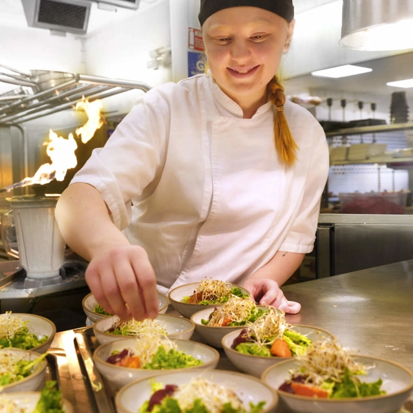 En kock lägger böngroddar på flera salladsskålar. Bakom henne använder en till kock en brännare för att förkolna något.