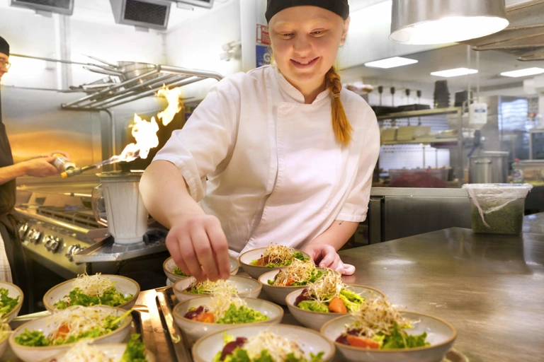 En kock lägger böngroddar på flera salladsskålar. Bakom henne använder en till kock en brännare för att förkolna något.