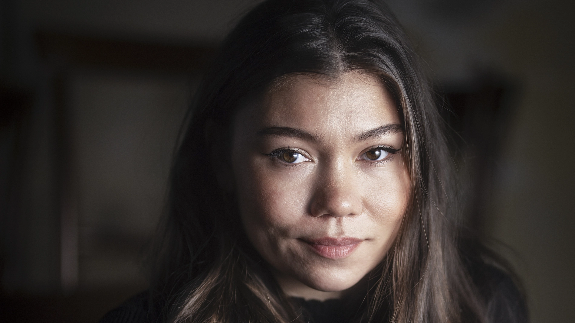 En musikartist sitter i ett mörkt rum och tittar in i kameran med neutralt ansiktsuttryck. Hon är belyst från vänster sida.