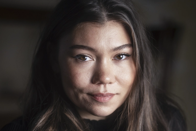 En musikartist sitter i ett mörkt rum och tittar in i kameran med neutralt ansiktsuttryck. Hon är belyst från vänster sida.