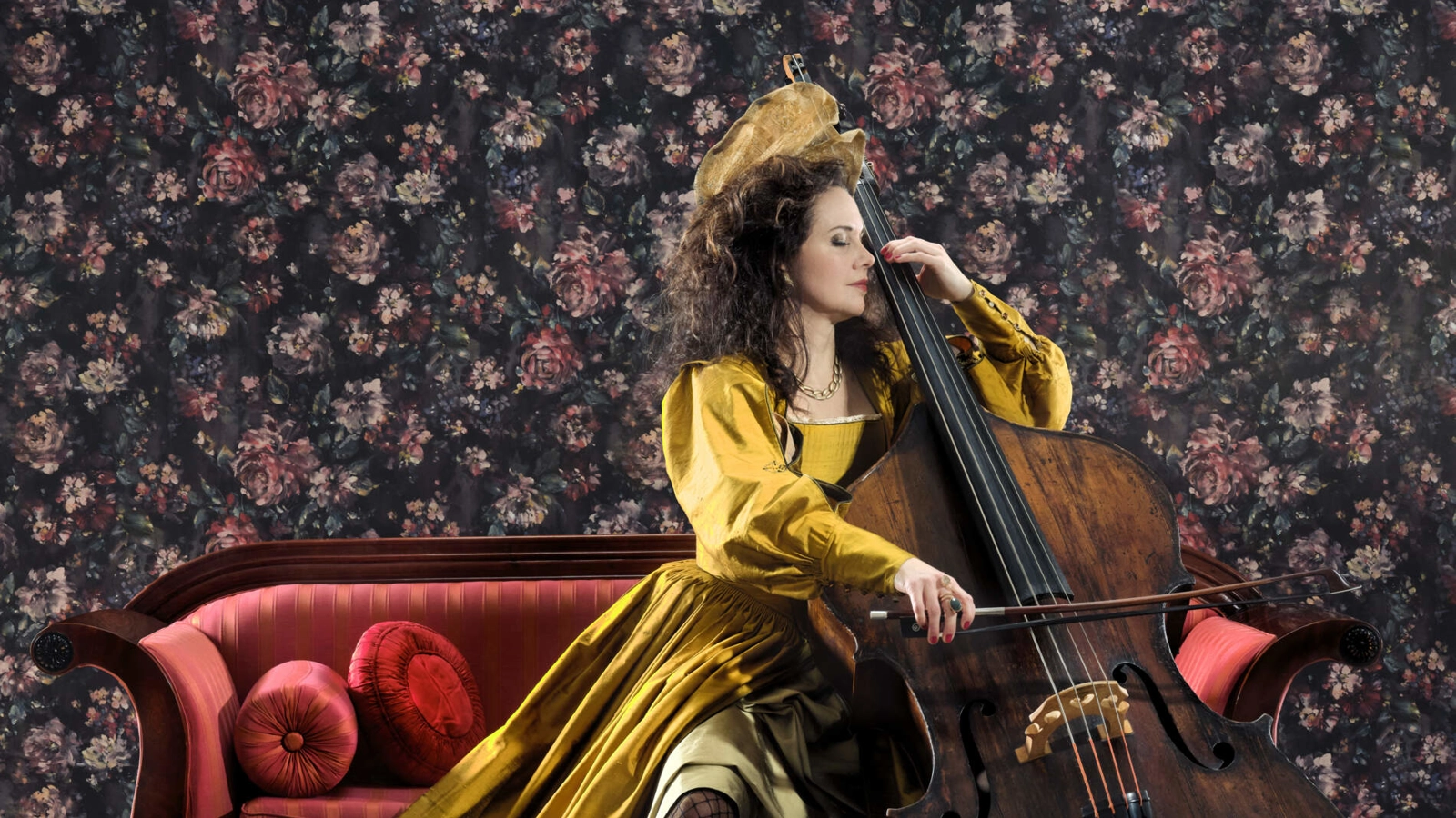 En cellist i gul sidenklänning sitter på en röd schäslong och spelar cellon. Bakom henne hänger en blommig bakgrund.