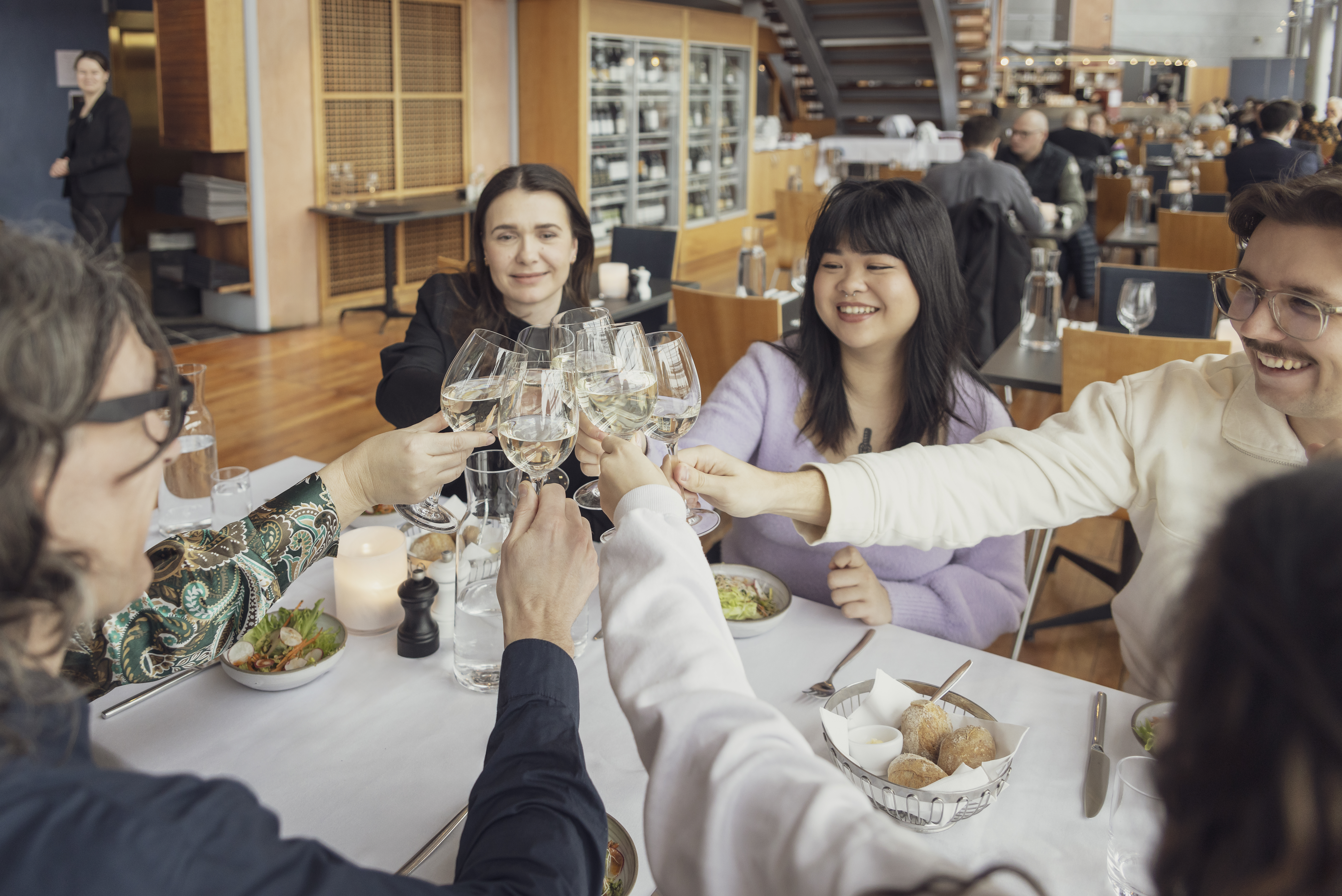 Fem personer vid ett restaurangbord håller ihop sina vinglas, ler och skålar.