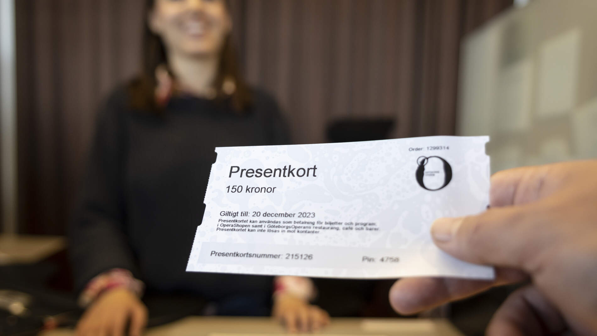 Närbild på en hand som räcker fram ett presentkort till ett leende person bakom disken i en biljettkassa.