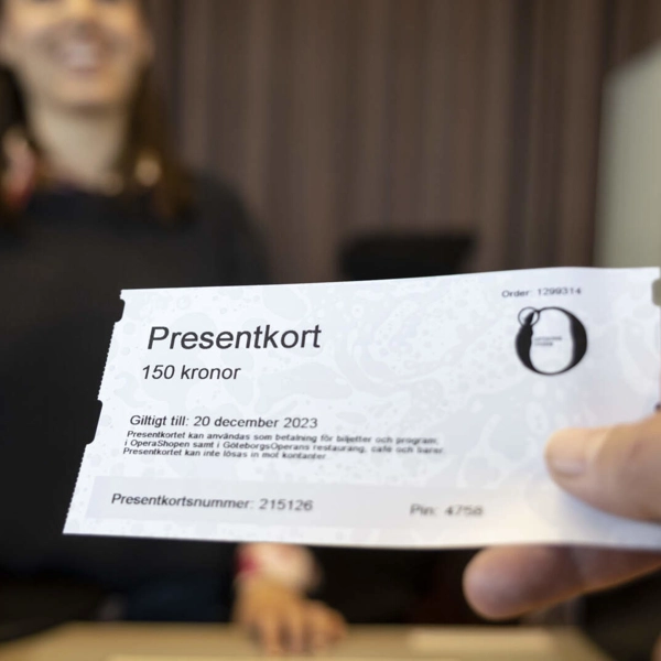 Närbild på en hand som räcker fram ett presentkort till ett leende person bakom disken i en biljettkassa.