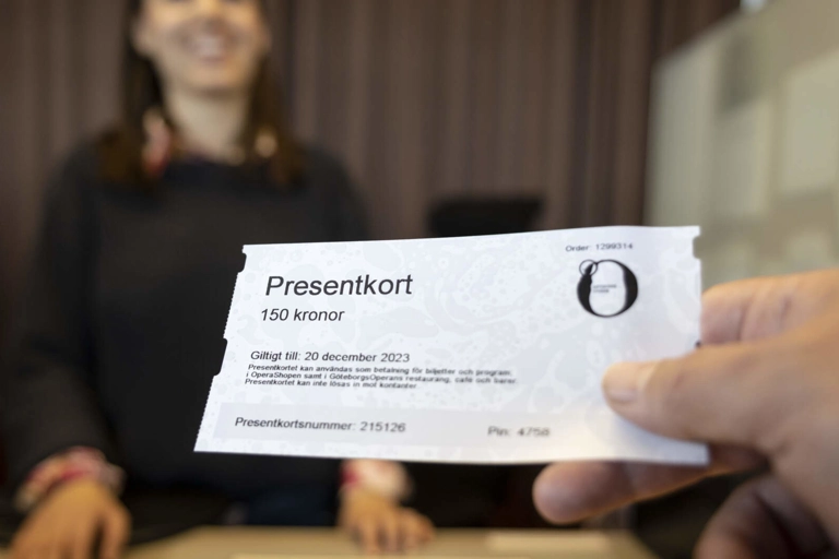 Närbild på en hand som räcker fram ett presentkort till ett leende person bakom disken i en biljettkassa.
