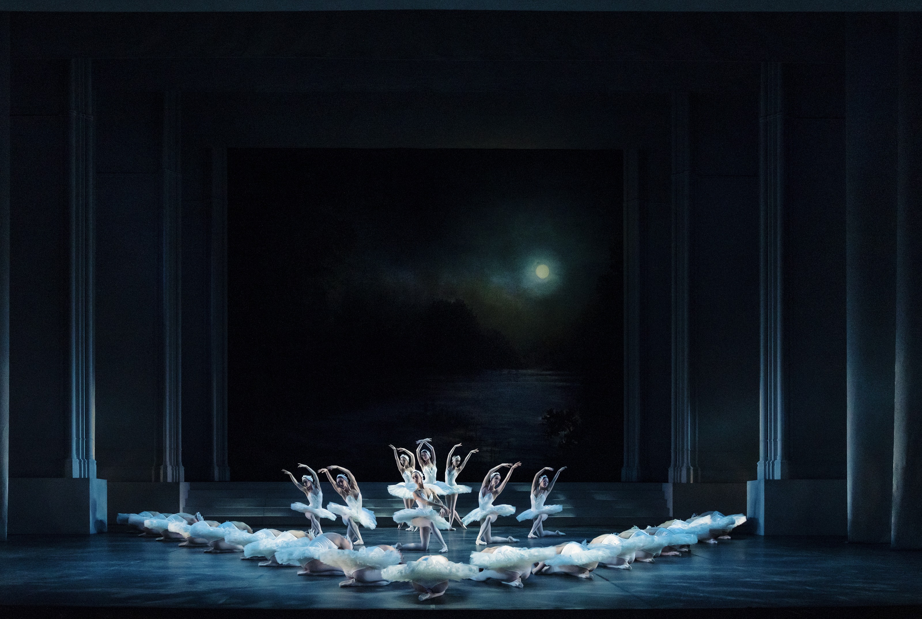 En grupp vitklädda ballerinor dansar synkroniserat på en mörk scen som svanar på en sjö. En fullmåne lyser bakom dem.