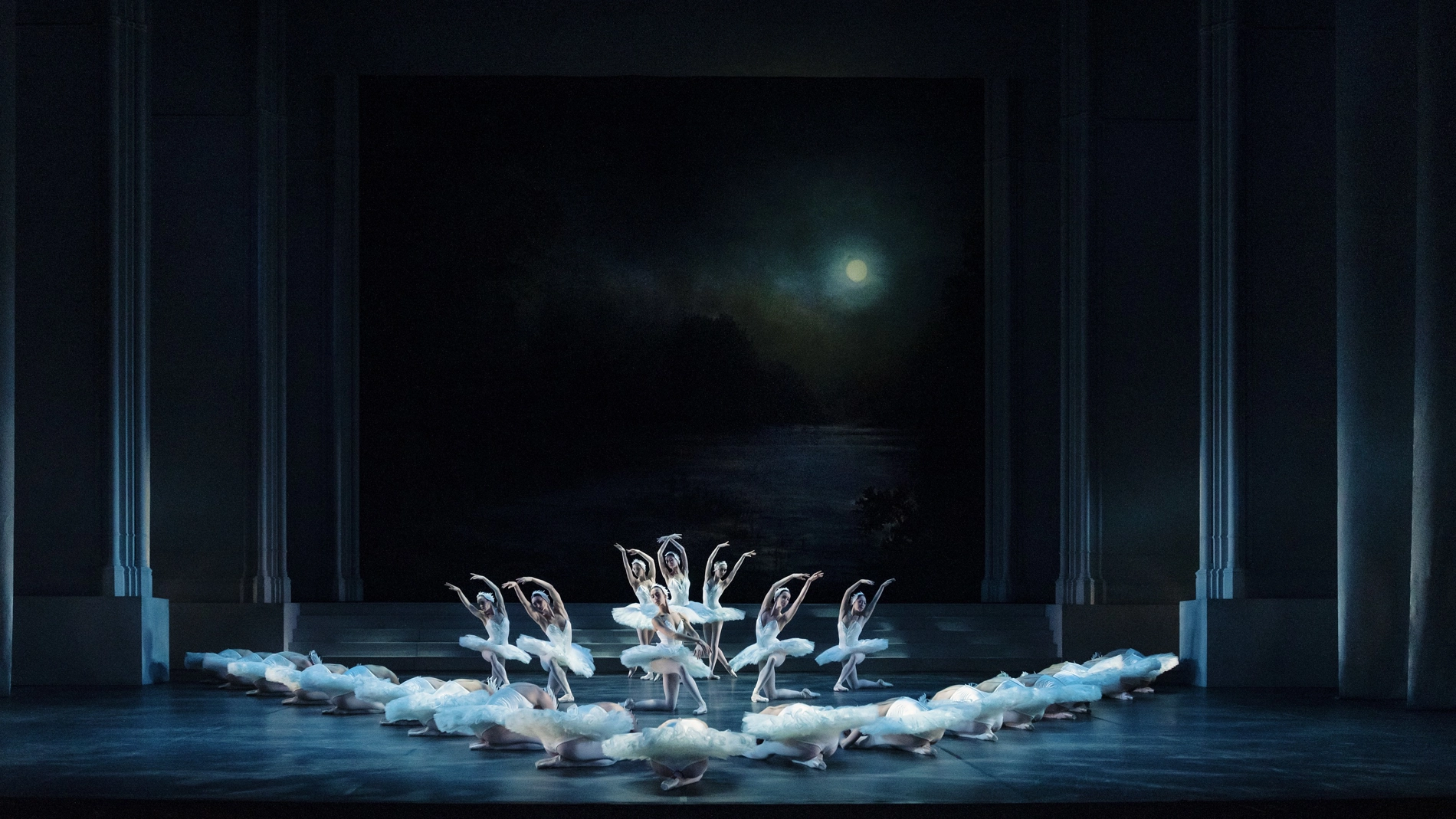 En grupp vitklädda ballerinor dansar synkroniserat på en mörk scen som svanar på en sjö. En fullmåne lyser bakom dem.